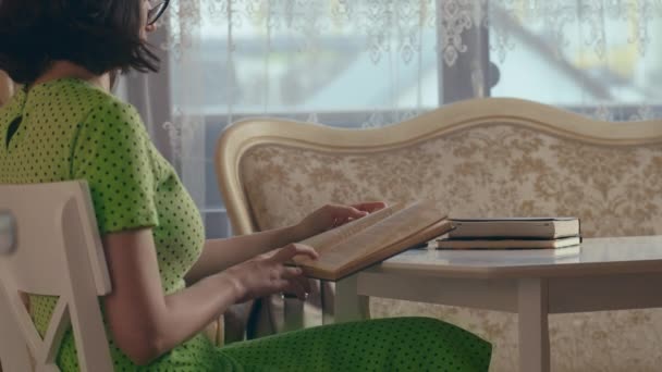 Una chica con un vestido verde sentada en una mesa leyendo un libro viejo, vista lateral — Vídeo de stock