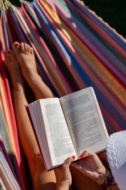 woman in white hat reading relaxed in a colourful hammock in the garden 
