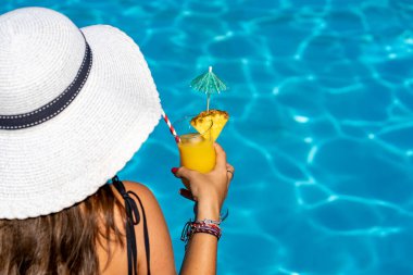 Young Caucasian woman in white hat enjoying a cocktail while sitting at the edge of the pool