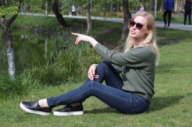 Yeşil kazaklı sarışın kız çimlerin üzerinde oturuyor.