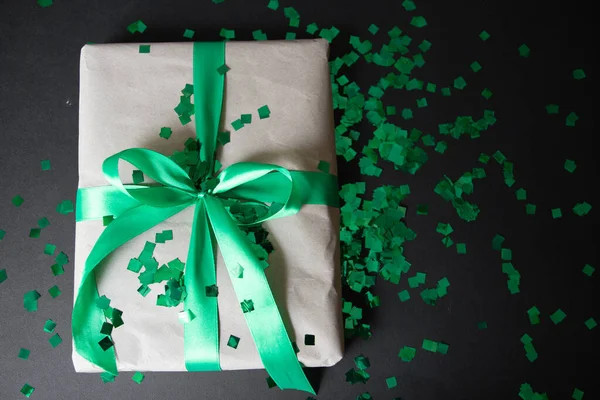 a gift in craft paper and a green ribbon on a black background with confetti