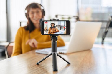 Kulaklıklardaki kadın radyo sunucusu stüdyoda mikrofon ve dizüstü bilgisayar kullanarak canlı yayın yapıyor. Kadın youtuber kameraya bakıyor, video çekiyor. Akıllı telefon kamerasına odaklan. Yakın plan.