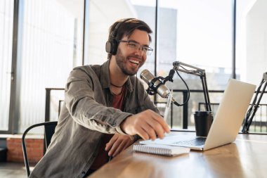 Kulaklık ve gözlüklü genç adam stüdyoda podcastın keyfini çıkarıyor, mikrofona konuşuyor, elinde kalem tutuyor, dizüstü bilgisayar kullanıyor. Yakışıklı podcaster canlı yayın yaparken gülüyor.