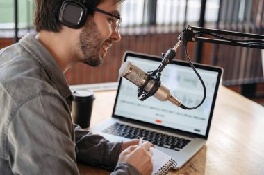 Erkek podcaster etkili blogcusu gülümserken kulaklıklar, dizüstü bilgisayarlar ve kulaklıklar kullanarak stüdyoda canlı yayın yapıyor. Gözlüklü erkek radyo sunucusu podcast veya röportaj yapıyor