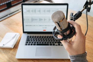 Yayın stüdyosunda mikrofon tutan adamın yakın çekimi. Ev stüdyosu podcast iç mekan. Dizüstü bilgisayar, kalem ve not defteri arka plandaki masada. Ev podcast stüdyosunda podcast yapıyorum. Birinci kişi görünümü