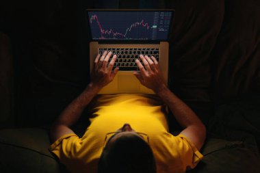 Genç işadamı yatırımcı bilgisayarındaki borsa uygulamasını evdeki kanepede kripto para birimi finansal piyasa analizi için kullanıyor, veri indeksi grafiği bilgisayar ekranında. Üst görünüm