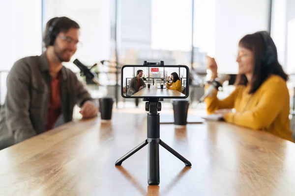 Kadın ve erkek stüdyoda canlı yayın yaparken birbirleriyle konuşuyorlar. Kulaklıklı iki podcaster tripoda akıllı telefon kullanarak canlı video çekiyor. Akıllı telefona seçici odaklanma