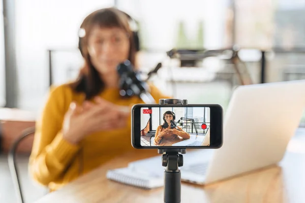 Dişi vlogger, mikrofon, dizüstü bilgisayar ve cep telefonunu kullanarak canlı yayın yapıyor. Kadın podcaster kayıt ve canlı yayın yapan akıllı telefon kamera ekranına seçmeli olarak odaklan. Yakın plan.