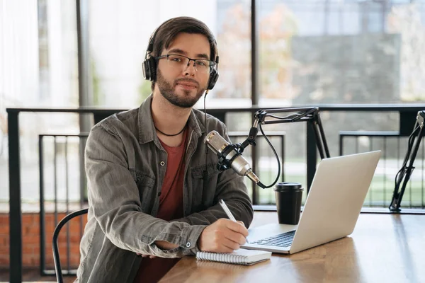 一个快乐的年轻人在录音棚里主持录制播客的肖像 一个面带微笑的英俊男人 戴着耳机和眼镜 带着笔记本电脑和话筒坐在桌旁 看着相机 用笔和笔记本 — 图库照片
