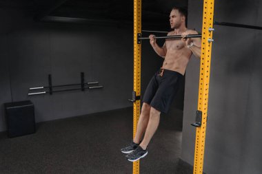 Kaslı üstsüz bir adam spor kulübündeki yatay barda barfiks çekiyor. Güçlü adam karanlık spor salonundaki barfiks çekiyor. Fonksiyonel eğitim, Crossfit ve fitness konsepti