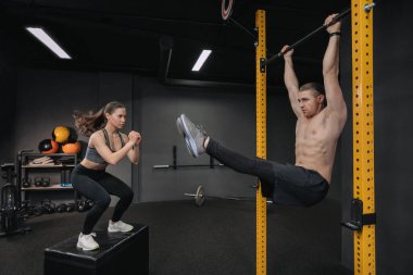 Crossfit eğitimi. Yakışıklı, üstsüz, kaslı bir adam karın egzersizi yaparken formda bir kadın atlama egzersizi yapıyor. Fonksiyonel antrenman, grup devresi eğitimi, fitness sınıfı.