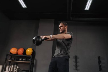 Spor salonunda kettlebell salıncakları yapan güçlü kaslı bir sporcu. Gri spor salonunun arka planında ekipmanla çekim yapmış. Halterci içeride egzersiz yapıyor, egzersiz yapıyor. Crossfit eğitmeni ısınıyor