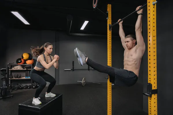 Crossfit eğitimi. Yakışıklı, üstsüz, kaslı bir adam karın egzersizi yaparken formda bir kadın atlama egzersizi yapıyor. Fonksiyonel antrenman, grup devresi eğitimi, fitness sınıfı.