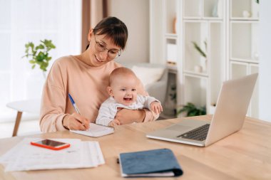 Neşeli bir iş kadını bebeğini kucağında tutuyor ve uzaktan ofisinde çalışıyor. Mutlu bekar anne gülümsüyor, not alıyor, online dersi var. Genç kadın serbest çalışan iş ve anneliği dengeliyor.