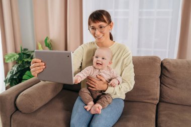 Tablette bebeğini elinde tutan mutlu bir anne. Genç bir kadın, gülümseyen oğluyla birlikte kanepede oturuyor. Kocasına veya akrabalarına çevrimiçi video araması yapmak için tablet kullanıyor. Çevrimiçi bağlantı