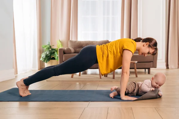 Beautiful yogi mom smiling at her baby while doing push up on exercising mat at home. Happy mom working out with her son. Young mom looking at her little baby during her post-natal fitness routine
