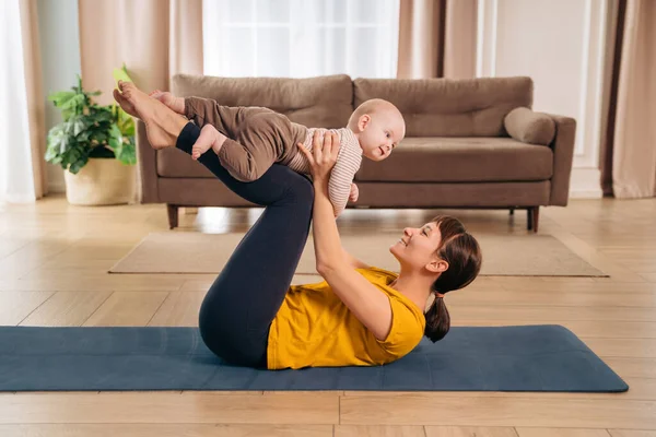 Yeni anne küçük bebeğini bacaklarıyla kaldırıyor, doğum sonrası fitness egzersizi yapıyor. Spor annesi evde bebekle fitness ve yoga yapıyor. Sağlıklı annelik ve doğum sonrası iyileşme