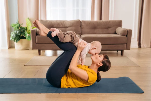 Bebekle spor. Anne, bebeğini kucağında tutarken ve evde spor paspası üzerinde doğum sonrası iyileşme egzersizi yaparken öpmeye hazır.