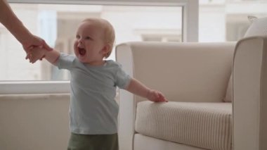 Neşeli erkek bebek oturma odasında ilk adımlarını atmaya çalışıyor. Atış becerisini geliştiriyor. Mutlu çocukluğunun tadını çıkaran meraklı gülen bir bebek. Anne küçük oğluna yürümeyi öğretiyor. Yavaş Hareket
