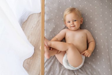 Upper view of sweet cute blue-eyed baby in diaper lying on his back in crib looking at camera, playing with foot putting legs up. Childcare concept. Happy carefree childhood. Copy space