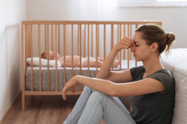 Side view of unhappy frustrated young mother sitting on floor in child bedroom while baby sleeping in bed, trying to calm down, suffering postnatal depression symptoms