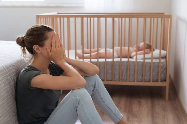 Side view of mother covering face crying from exhaustion sitting on floor next to baby crib while child napping, cant handle difficulties of maternity, having postnatal depression symptoms