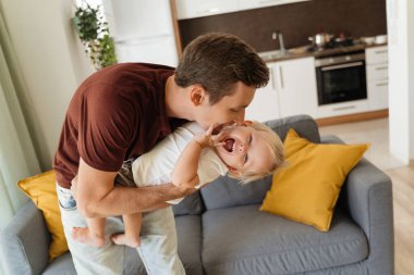 Young father cuddling his baby son after returning home from work, hugging, tickling holding him in hands on kitchen background and gray sofa in living-room. Happy carefree childhood, fatherhood