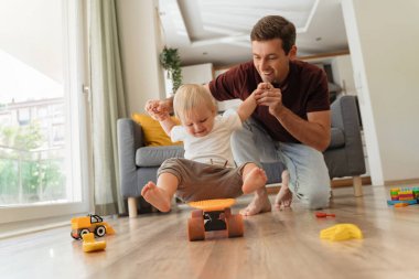 Little cute baby boy sitting and riding skateboard while his father holding his hands, having fun playing together in living-room after lunch and nap time. Carefree childhood, happy fatherhood