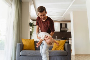 Young father spending time with his baby boy, cuddling and playing active games in living-room after returning home from work, carrying child as airplane, swaying together on background with gray sofa