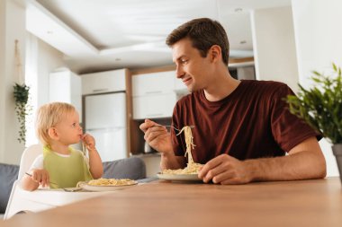 Side view of happy father and baby boy having spaghetti on dinner sitting at kitchen table, son in bib in high chair learning to eat with fork. Self-feeding, weaning