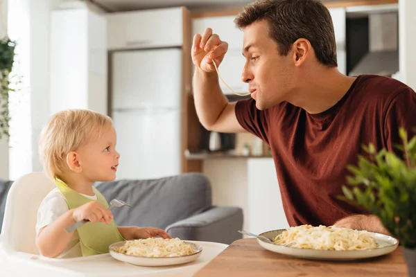 Happy dad playing with spaghetti while having dinner with his baby boy in bib learning to eat with fork looking at funny dad with interest. Happy fatherhood, feeding kid. Father day, family