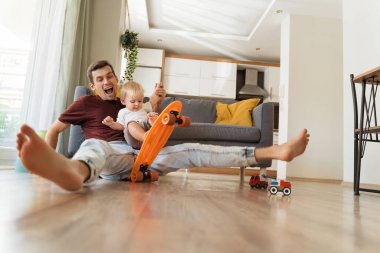 Funny image of happy cheerful baby boy spending time with his caring dad or brother sitting on floor wheeling skateboard, pretending like falling down, father entertaining his son at home. Fatherhood