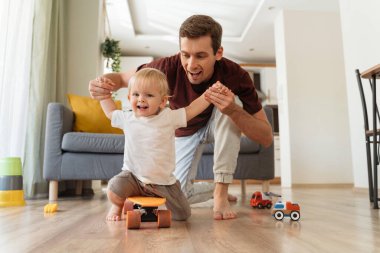 Happy young daddy holding his baby boys hands while cute little blond toddler sitting and wheeling on skateboard in living-room at home with couch on background. Fatherhood, happy childhood