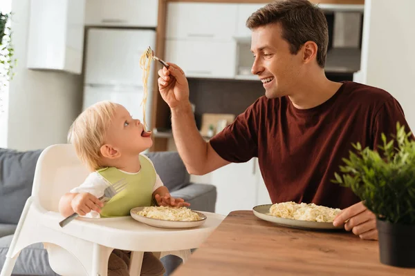 Happy fathers day. Side view of dad feeding baby in green bib with spaghetti during lunch time at kitchen table, kid sitting in high chair holding fork. Happy fatherhood, first meal, self-feeding