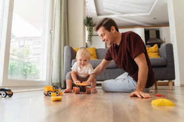 Cute blond baby boy sitting on orange skateboard on floor in living-room playing with daddy in active games and developmental toys at home. Happy carefree childhood. Father spending time with his son