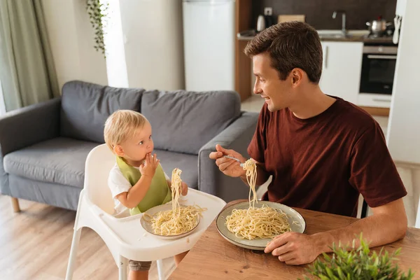 Upper view of handsome dad and cute blond baby in high chair eating spaghetti for lunch at kitchen table, little kid learning to eat with fork. Happy fatherhood. Father feeding his son, having fun