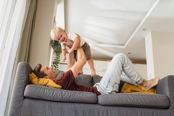Happy dad playing with his toddler, lifting him up lying on sofa in living room, having fun together, enjoying weekend. Carefree childhood, father and son relationships. Active play