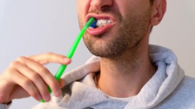 Diş teli takan beyaz bir adam arka planda diş fırçasıyla dişlerini fırçalar.