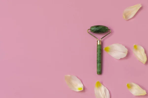 Roller wajah terbuat dari batu giok hijau alami di latar belakang merah muda dengan kelopak putih — Stok Foto