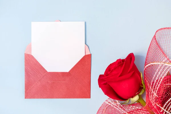 Top view of red envelope and white empty card with beautiful rose on light blue background, copy space