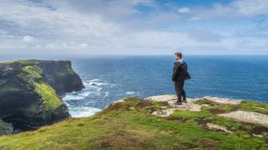 Moher 'in ikonik uçurumlarının tepesinde duran ve muhteşem manzaraya hayran olan adam, popüler turistik ilgi, Vahşi Atlantik Yolu, County Clare, İrlanda