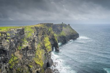 Turistler, Moher Kulesi ile uzak mesafede İrlanda 'da Moher Kayalıklarını geziyorlar.