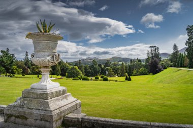 An ornate pot with flowers, wide green lawn with bushes in Powerscourt gardens, forest and Sugerloaf mountain in the background, Wicklow, Ireland