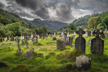 Wicklow, İrlanda, Ağustos 2019 Glendalough Mezarlığı 'nda Kelt haçları olan antik mezarlar dağlar ve arka planda dramatik fırtına gökyüzü