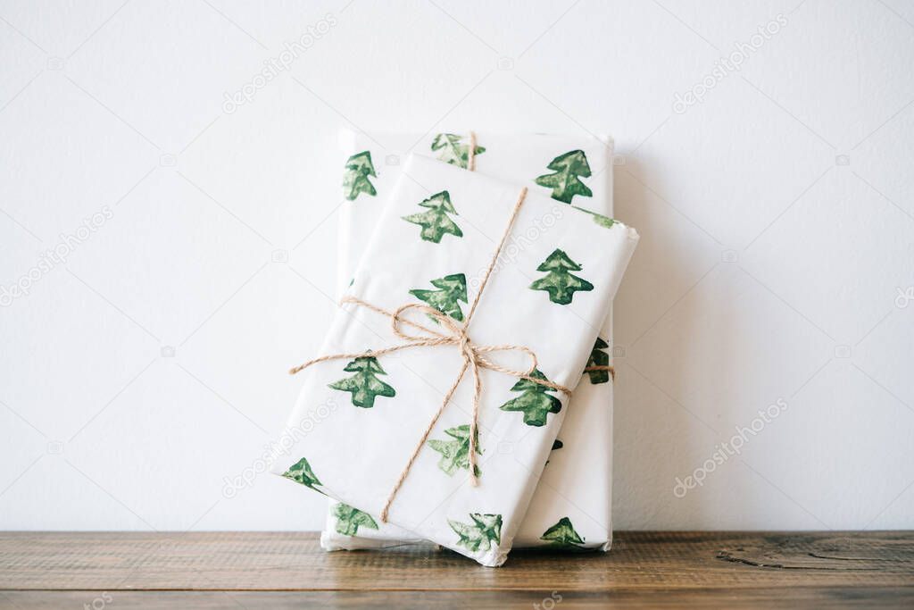 the New Year's gift is packed in festive paper with watercolor Christmas trees on a wooden table against a white wall. High quality photo