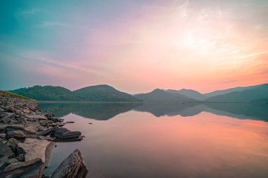 Güneş doğarken yansıması mükemmel olan dağ gölü manzarası. Pembe pastel gökyüzü, arka planda tepeler ve suya yansıyan güzel dağ manzarası. Doğa gölü manzarası