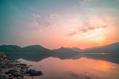Güneş doğarken yansıması mükemmel olan dağ gölü manzarası. Pembe pastel gökyüzü, arka planda tepeler ve suya yansıyan güzel dağ manzarası. Doğa gölü manzarası