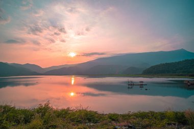 Güneş doğarken yansıması mükemmel olan dağ gölü manzarası. Pembe pastel gökyüzü, arka planda tepeler ve suya yansıyan güzel dağ manzarası. Doğa gölü manzarası