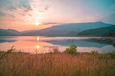 Güneş doğarken yansıması mükemmel olan dağ gölü manzarası. Pembe pastel gökyüzü, arka planda tepeler ve suya yansıyan güzel dağ manzarası. Doğa gölü manzarası