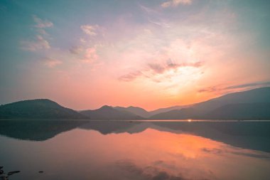 Güneş doğarken yansıması mükemmel olan dağ gölü manzarası. Pembe pastel gökyüzü, arka planda tepeler ve suya yansıyan güzel dağ manzarası. Doğa gölü manzarası